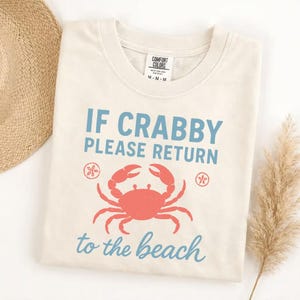 May include: Cream-colored t-shirt with the text "IF CRABBY PLEASE RETURN to the beach" in blue script. A coral-colored crab graphic is centered below the text. The shirt is a Comfort Colors brand.