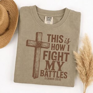 May include: A sage green t-shirt with the text "THIS is HOW I FIGHT MY BATTLES" and a brown cross graphic. The shirt also has the Bible verse reference "2 CHRON 20:15". A straw hat and dried grass are in the background.