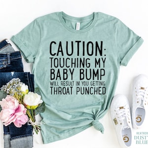 Könnte beinhalten: Ein hellblaues T-Shirt mit dem Text "CAUTION: TOUCHING MY BABY BUMP WILL RESULT IN YOU GETTING THROAT PUNCHED" in Schwarz gedruckt.