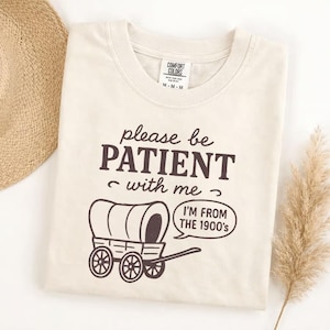 May include: Cream-colored t-shirt with the text "please be PATIENT with me" and a covered wagon graphic. The phrase "I'M FROM THE 1900's" is in a speech bubble. The shirt has a crew neck and short sleeves.