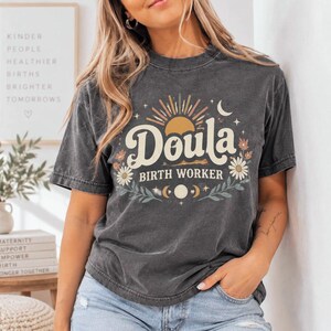Puede incluir: Una camiseta gris carbón de manga corta con un estampado gráfico que incluye el texto Doula Birth Worker. El diseño presenta un sol, una luna, estrellas y elementos florales en colores crema, naranja y verde en el frente.
