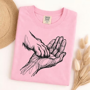 May include: A light pink Comfort Colors t-shirt featuring a black line art illustration of two hands. One hand is cupping the other. The shirt has a crew neck and a tag that reads "Comfort Colors".