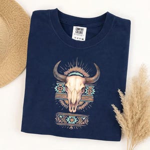 May include: A navy blue t-shirt featuring a watercolor design of a bull skull with large horns. The design includes geometric patterns in shades of turquoise, orange, and brown. The shirt has a crew neck and the brand name "COMFORT COLORS" is visible.