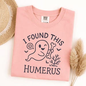 May include: A light pink Comfort Colors sweatshirt with the phrase "I FOUND THIS HUMERUS" in dark blue. The graphic features a cartoon ghost holding a bone, with a skull speech bubble and bats.