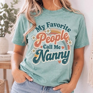 Puede incluir: Una camiseta de manga corta en color verde espuma de mar con un estampado gráfico que dice My Favorite People Call Me Nanny. El diseño incluye un arcoíris multicolor, pequeñas margaritas blancas y corazones rojos de estilo retro.