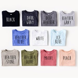 May include: A variety of folded t-shirts in different colors, including black, dark gray, deep heather gray, heather gray, heather blue, white, ice blue, heather stone, heather olive, mauve, and peach. Each shirt has a different color and text printed on it.