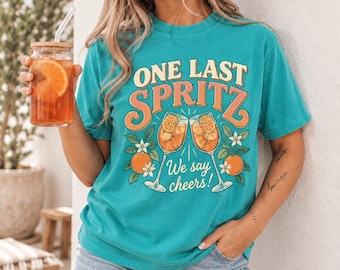 May include: A teal short-sleeve t-shirt featuring a graphic print of two clinking cocktail glasses filled with orange slices. The text reads ONE LAST SPRITZ We say cheers! in orange and white lettering with orange and green botanical accents.