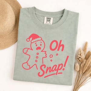 May include: A sage green t-shirt with a red graphic of a gingerbread man wearing a Santa hat and the words "Oh Snap!". The shirt is made of a soft material and has a crew neck.