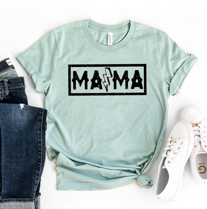 Pu&ograve; includere: Una maglietta blu chiaro con una grafica nera che dice "MAMA" con un fulmine al centro. La grafica ha un aspetto usurato.