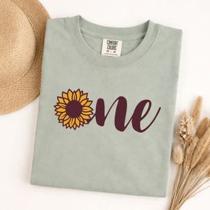 以下が含まれることがあります： 黒いハンドルと「But First, Coffee」のテキストが黒い太字で書かれた白いセラミックマグ。マグは白い表面に置かれています。