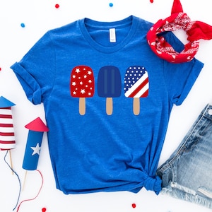 May include: A blue t-shirt with a graphic of three popsicles. The popsicles are decorated with red, white, and blue stars and stripes.