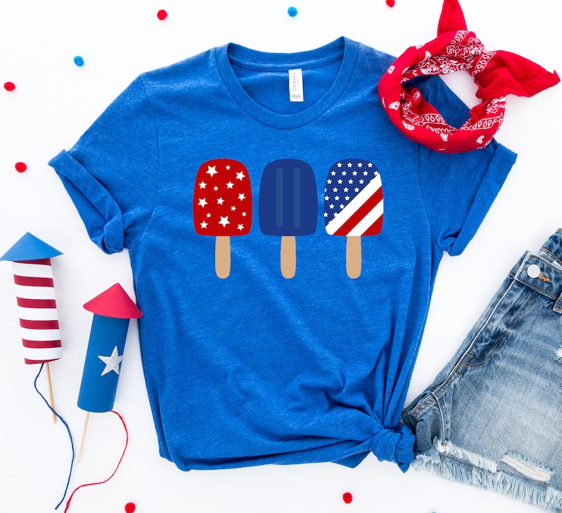 May include: A blue t-shirt with a graphic of three popsicles. The popsicles are decorated with red, white, and blue stars and stripes.