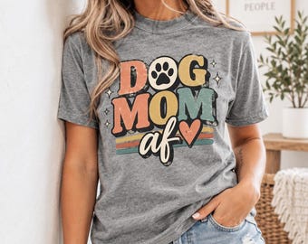 May include: A heather gray short-sleeve t-shirt featuring the text DOG MOM af in a multi-colored retro font. The design includes a black paw print, a red heart, and horizontal stripes in muted orange, teal, and red, accented by small stars.
