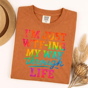 May include: A rust-colored t-shirt with the text "I'M JUST WTF-ING MY WAY THROUGH LIFE" in rainbow-colored letters. The shirt is folded and has a tag that says "Comfort Colors". A straw hat and dried flowers are in the background.