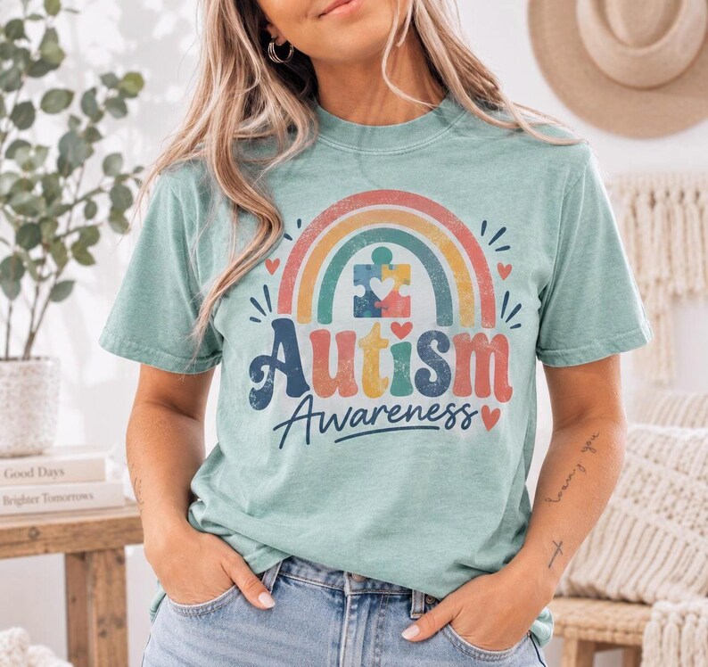 A woman wearing a light blue t-shirt with a colorful graphic design that says "Autism Awareness" and a rainbow symbol.
