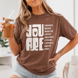 May include: A brown short-sleeve t-shirt featuring white text that reads YOU ARE with a heart icon, followed by a list of words: ENOUGH, BRAVE, WORTHY, LOVED, AMAZING, STRONG, CAPABLE, SMART, and KIND. The shirt has a crew neckline and a relaxed fit.