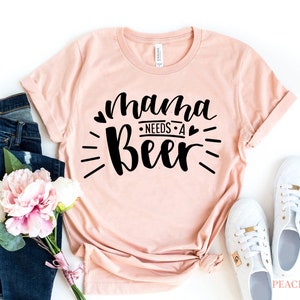 May include: A peach-colored t-shirt with the text "mama needs a beer" in black lettering with hearts and a sunburst design.