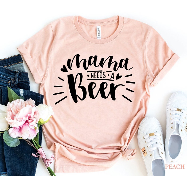 May include: A peach-colored t-shirt with the text "mama needs a beer" in black lettering with hearts and a sunburst design.