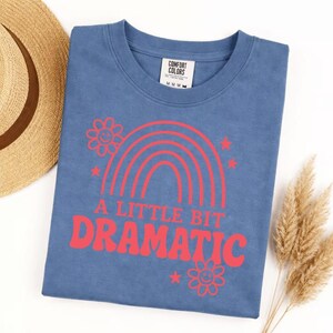 May include: A blue t-shirt with the text "A Little Bit Dramatic" in red, along with a red rainbow, stars, and flowers. The shirt is folded and lies on a white surface, with a straw hat and dried plants in the background.