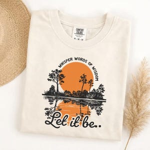 May include: Cream-colored t-shirt featuring a graphic of a sunset over a lake with trees. The text "Whisper Words of Wisdom" arches above the sun, and "Let it be.." is written below. The shirt has a crew neck and short sleeves.