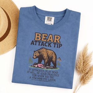 May include: A blue t-shirt with the text "BEAR ATTACK TIP" and a graphic of a bear and a person. The text below reads "IF ATTACKED, PLAY DEAD IT WILL BE GOOD PRACTICE FOR WHEN YOU DIE A FEW MINUTES LATER."