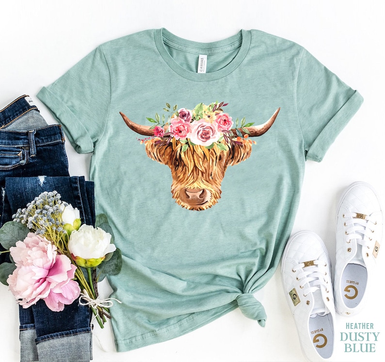 May include: A light blue t-shirt with a watercolor illustration of a Highland cow wearing a floral crown. The cow is brown and white with a long, shaggy mane. The flowers in the crown are pink, white, and yellow.