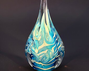 May include: A teardrop-shaped glass paperweight with a swirling blue and white design. The glass is clear and has small air bubbles trapped inside.
