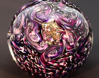 May include: A glass paperweight with a swirling pattern of purple, black, and white. The paperweight has small air bubbles throughout and a gold fleck in the center.