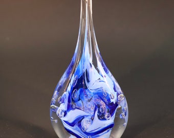 May include: A clear glass teardrop-shaped vase with a blue and white swirl design and silver glitter. The vase has small air bubbles trapped inside the glass.