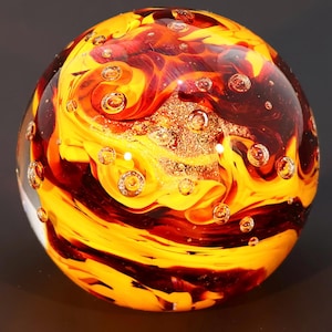 May include: A glass paperweight with a swirling pattern of red and yellow glass. The glass is filled with small air bubbles.