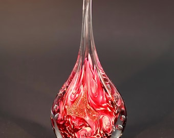 May include: A clear glass art piece with a teardrop shape. The interior features swirling red and white patterns with gold glitter accents. Small air bubbles are visible within the glass, and the piece has a long, slender, clear stem.