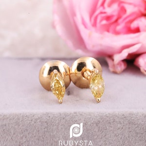 May include: A pair of gold stud earrings featuring marquise-cut yellow gemstones. The earrings have round gold backings and are positioned upright. The text RUBYSTA JEWELS is printed at the bottom with a circular logo above the brand name.