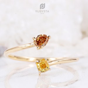 May include: A gold bypass ring featuring two distinct gemstones. One end holds a pear-shaped deep orange stone, while the other end features a square-cut bright yellow stone. The band is polished gold, and the logo RUBYSTA U.S.A. is visible.