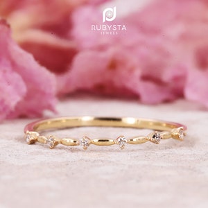 Op de afbeelding: Een dunne gouden ring met een reeks kleine, heldere, rond geslepen stenen die met tussenpozen langs de band zijn gezet, afgewisseld met gladde, langwerpige gouden metalen segmenten. De tekst RUBYSTA JEWELS staat in witte letters boven de ring.