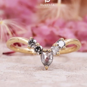 May include: A gold ring with a V-shaped design featuring five grey diamonds. The ring is set against a pink floral background.