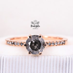 May include: A rose gold engagement ring with a black diamond center stone and a pave diamond band. The ring is in a white box with the text "Rubysta Jewels" on the top.