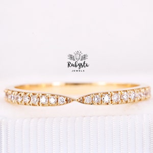 May include: A gold band ring featuring a curved, V-shaped center point and a row of small, clear, round-cut stones set along the entire length of the band. Above the ring, the text Rubysta JEWELS is printed with a small crystal icon.