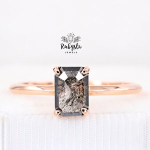 May include: A rose gold engagement ring with a black emerald cut diamond. The diamond has a unique, cloudy appearance.