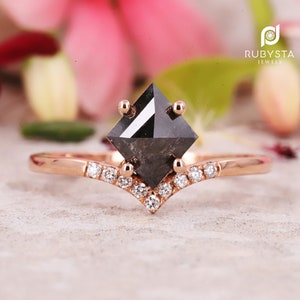 May include: A rose gold engagement ring with a black square diamond and a v-shaped band of smaller diamonds. The ring is set on a gray surface.