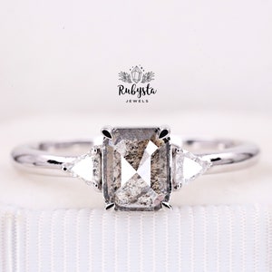 May include: A silver-toned ring featuring a rectangular salt and pepper diamond center stone held by four prongs, flanked by two smaller triangular side stones. The text Rubysta JEWELS is printed above the ring with a small crystal icon.