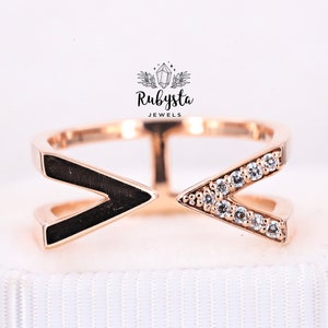 May include: A rose gold ring featuring a split band design with two V-shaped ends. One side is inlaid with a smooth black material, while the other side is set with a row of clear, sparkling stones. The logo Rubysta JEWELS is visible above.