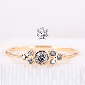 May include: A gold ring featuring a central round salt and pepper diamond in a bezel setting, flanked by six smaller round salt and pepper diamonds in a cluster arrangement. The text Rubysta JEWELS is printed above the ring in black ink.