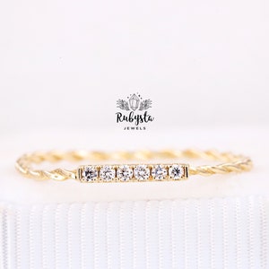 May include: A gold ring featuring a twisted band and a horizontal bar set with six small, clear, round-cut stones. Above the ring, the text Rubysta JEWELS is displayed with a small crystal icon. The ring is positioned against a white background.