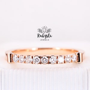 May include: A rose gold band ring featuring a row of ten round, clear, faceted stones set in a channel-style setting. Above the ring, the text Rubysta JEWELS is displayed with a small graphic of a crystal surrounded by leaves.