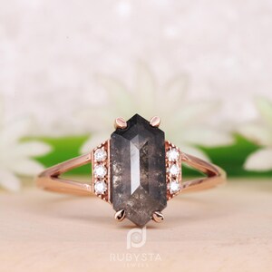 May include: A rose gold engagement ring with a hexagonal grey diamond and a halo of smaller diamonds.