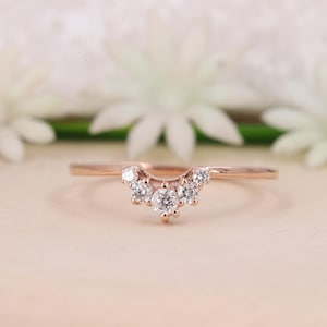 Peut inclure: Une bague en or rose dotée d'un anneau incurvé en forme de V, serti de cinq pierres rondes transparentes. La pierre centrale est la plus grande, flanquée de deux pierres plus petites de chaque côté, créant un agencement symétrique et gradué.