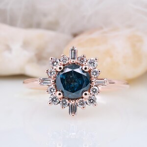 Peut inclure: Une bague de fiançailles en or rose avec une grande pierre centrale ronde de couleur bleu foncé entourée d'un halo de diamants plus petits. La bague a un design floral délicat.