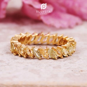 May include: A gold eternity band featuring a continuous row of marquise-cut yellow gemstones set in a shared prong setting. The ring displays a vibrant array of golden and light yellow hues, creating a cohesive and sparkling circular design.