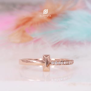 Peut inclure: Une bague en or rose avec un motif central en forme de T. Un côté de l'anneau est lisse, tandis que l'autre côté est serti d'une rangée de petites pierres claires taillées en rond. Le logo RUBYSTA USA est visible au-dessus de la bague.