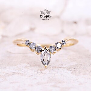 Peut inclure: Une bague dorée avec un anneau incurvé, ornée d'une pierre centrale grise de forme marquise et de petites pierres grises rondes. La bague est posée sur une surface claire et texturée. La marque "Rubysta Jewels" est visible.